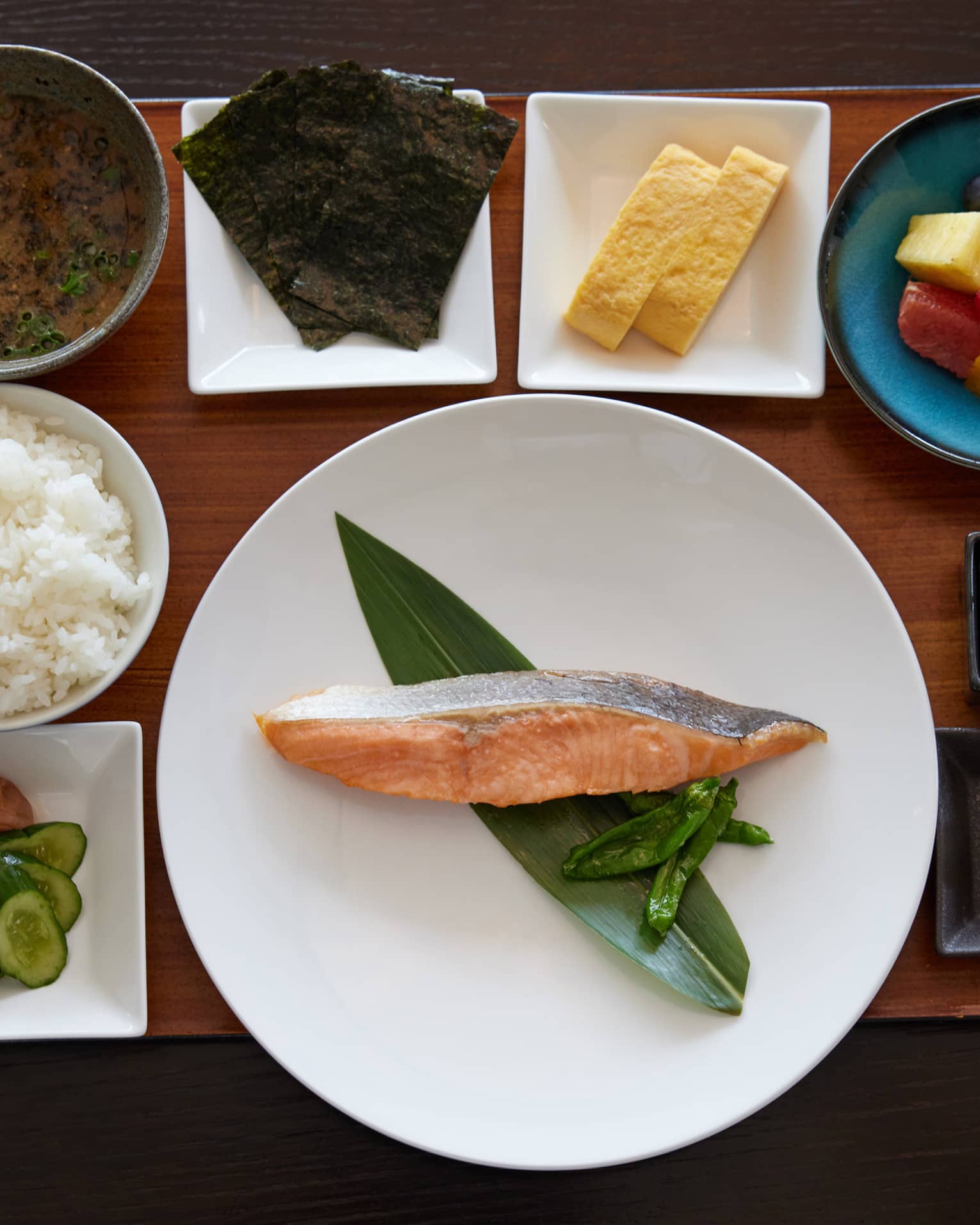 On wooden tray in dishes of various shapes and sizes, an alluring spread of food items, including fish, rice and fresh fruit.