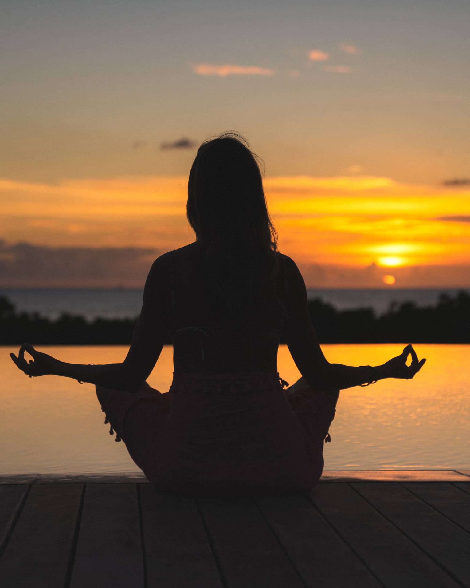 Silhouette of woman in meditation pose in front of calm ocean and orange sunset sky
