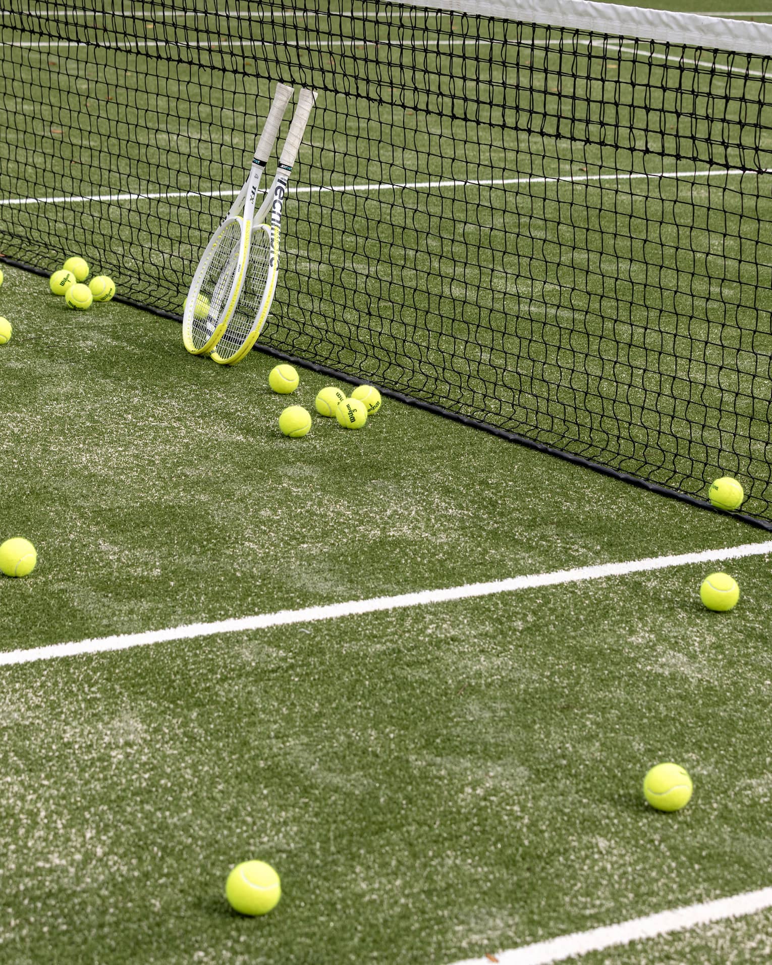 Tennis balls scattered on a tennis court. Two tennis racquets are propped against the net.