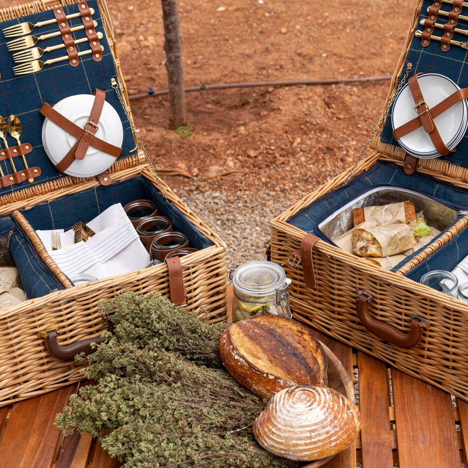 Plates and gold cutlery secured to the lids of picnic baskets containing parchment-wrapped sandwiches, napkins, and glasses.