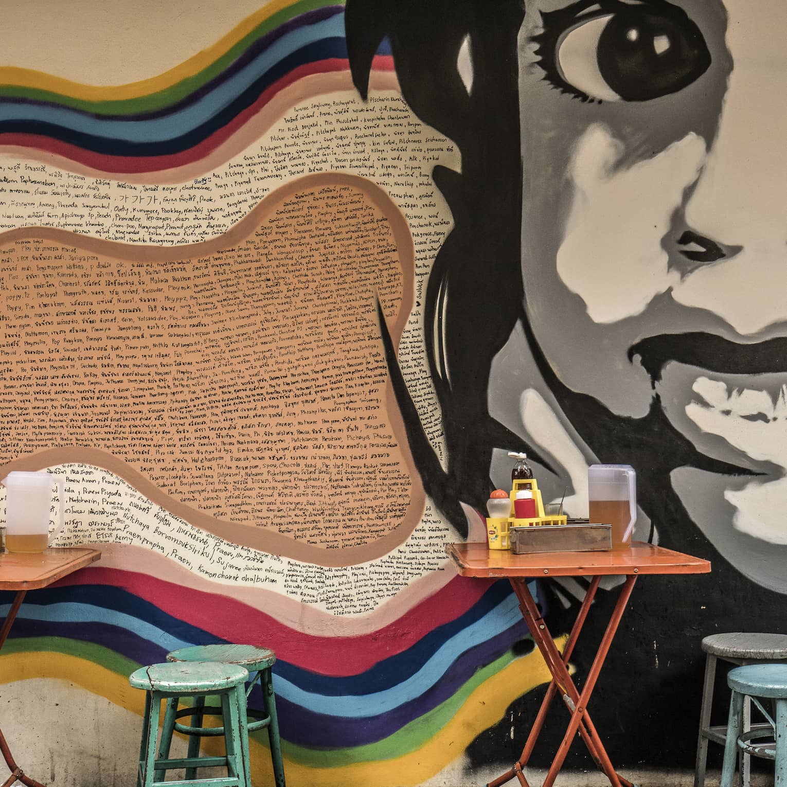 Street art mural featuring a large black-and-white face on a wall, with small, colourful dining tables and stools in front it