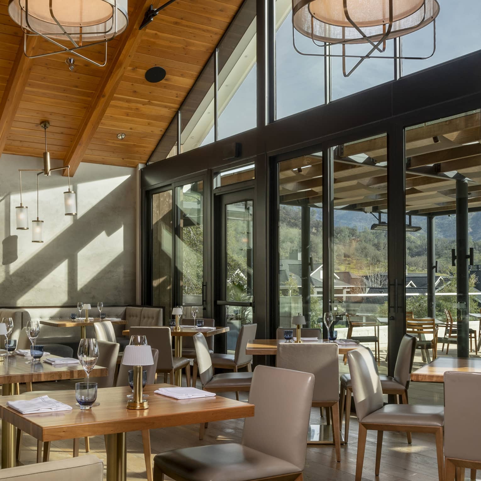 Modern restaurant interior with high wooden ceilings, large windows, and natural light. Tables are set, and an outdoor terrace offers views of surrounding hills.