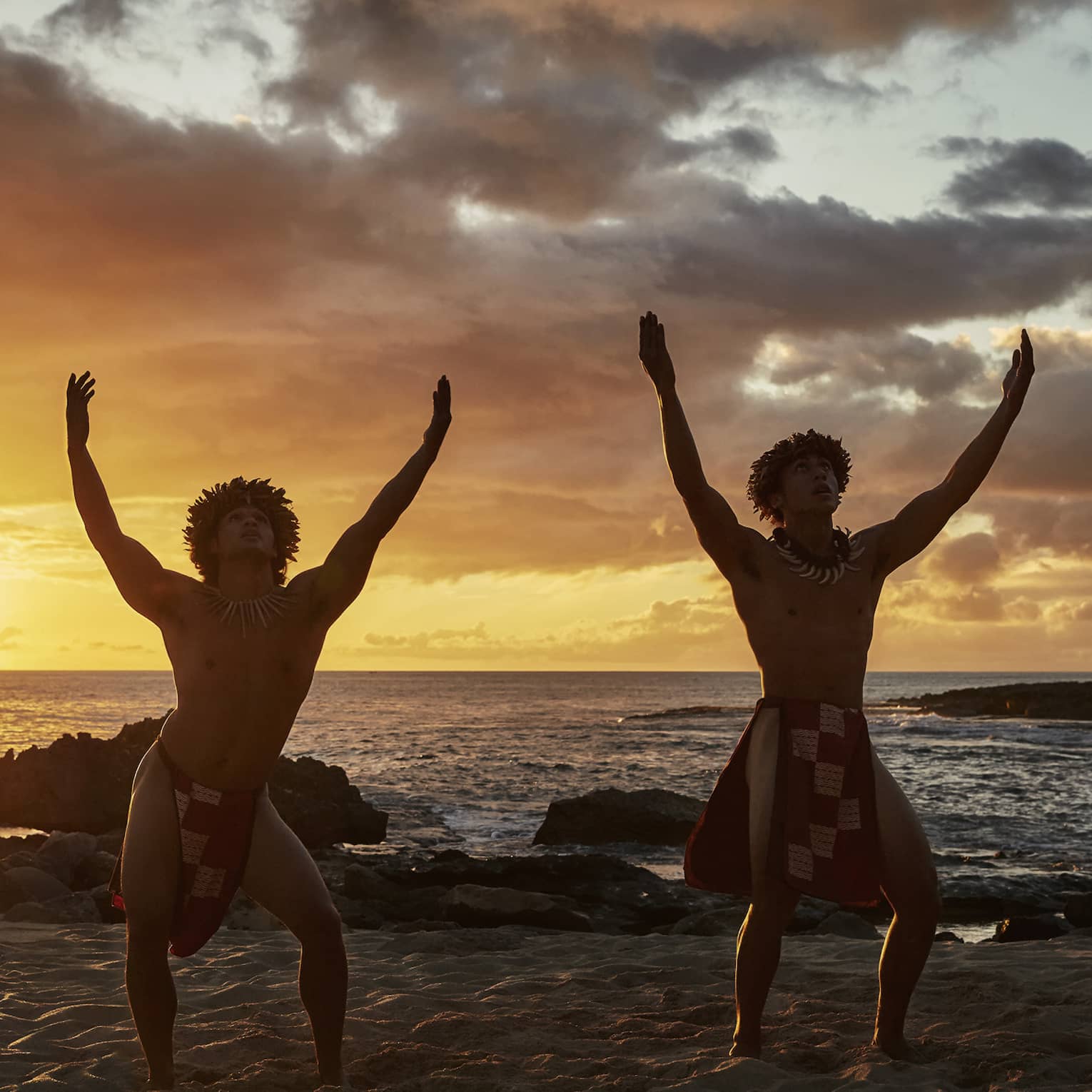 Two hula dancers on the beach, sinewy arms raised above their heads, in silhouette against dramatic fiery clouds at sunset.