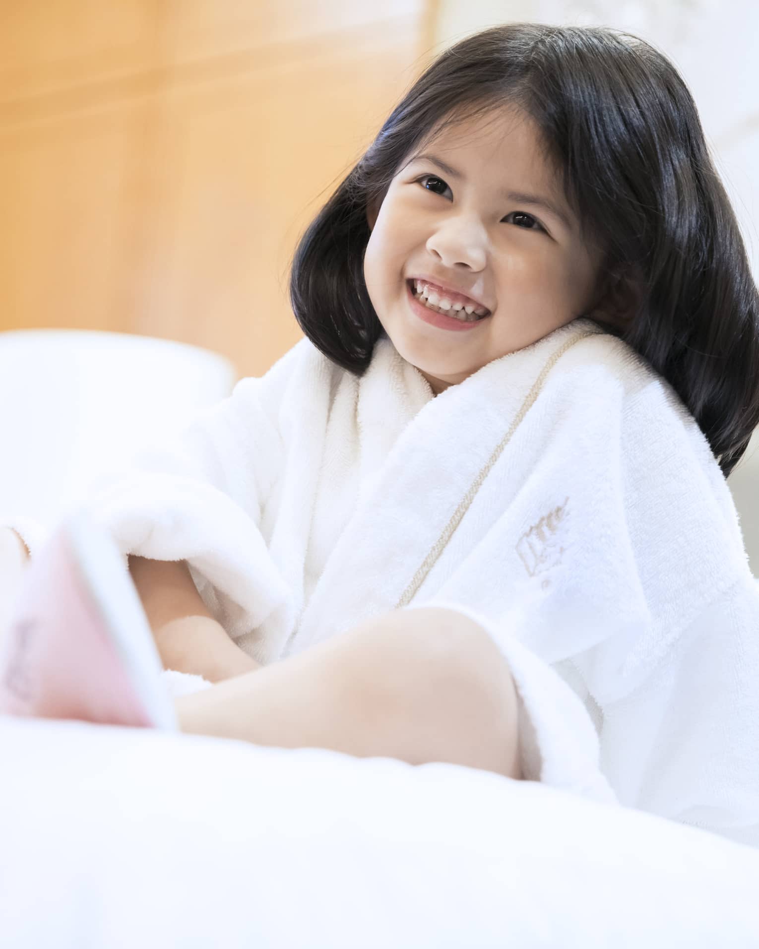 Smiling little girl wearing white bath robe and pink slippers sits on bed