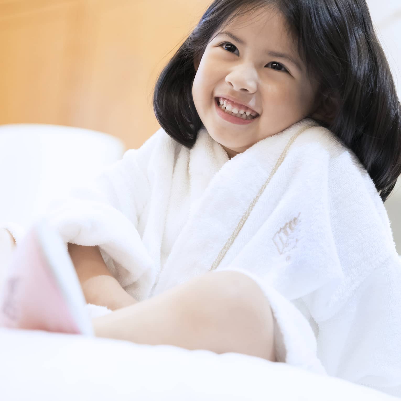 Smiling little girl wearing white bath robe and pink slippers sits on bed