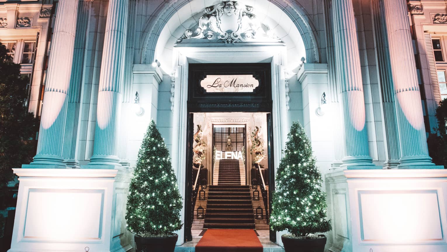 A Four Seasons Hotel Entrance is festively decorated with clear christmas lights on two small evergreen bushes