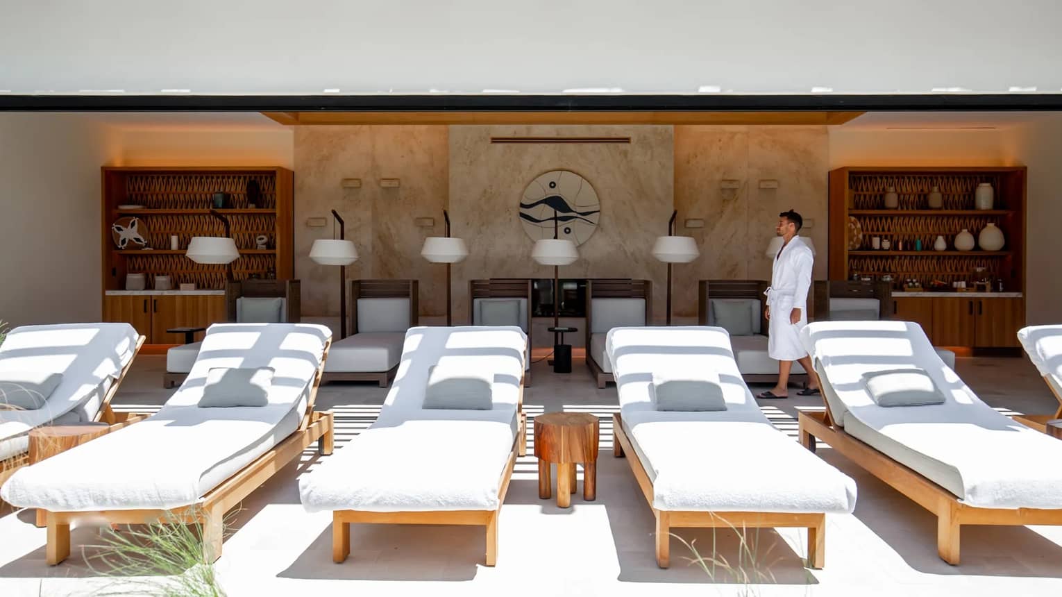 A man dressed in a white spa robe walks in an outdoor spa pavillion behind a row of white lounge chairs