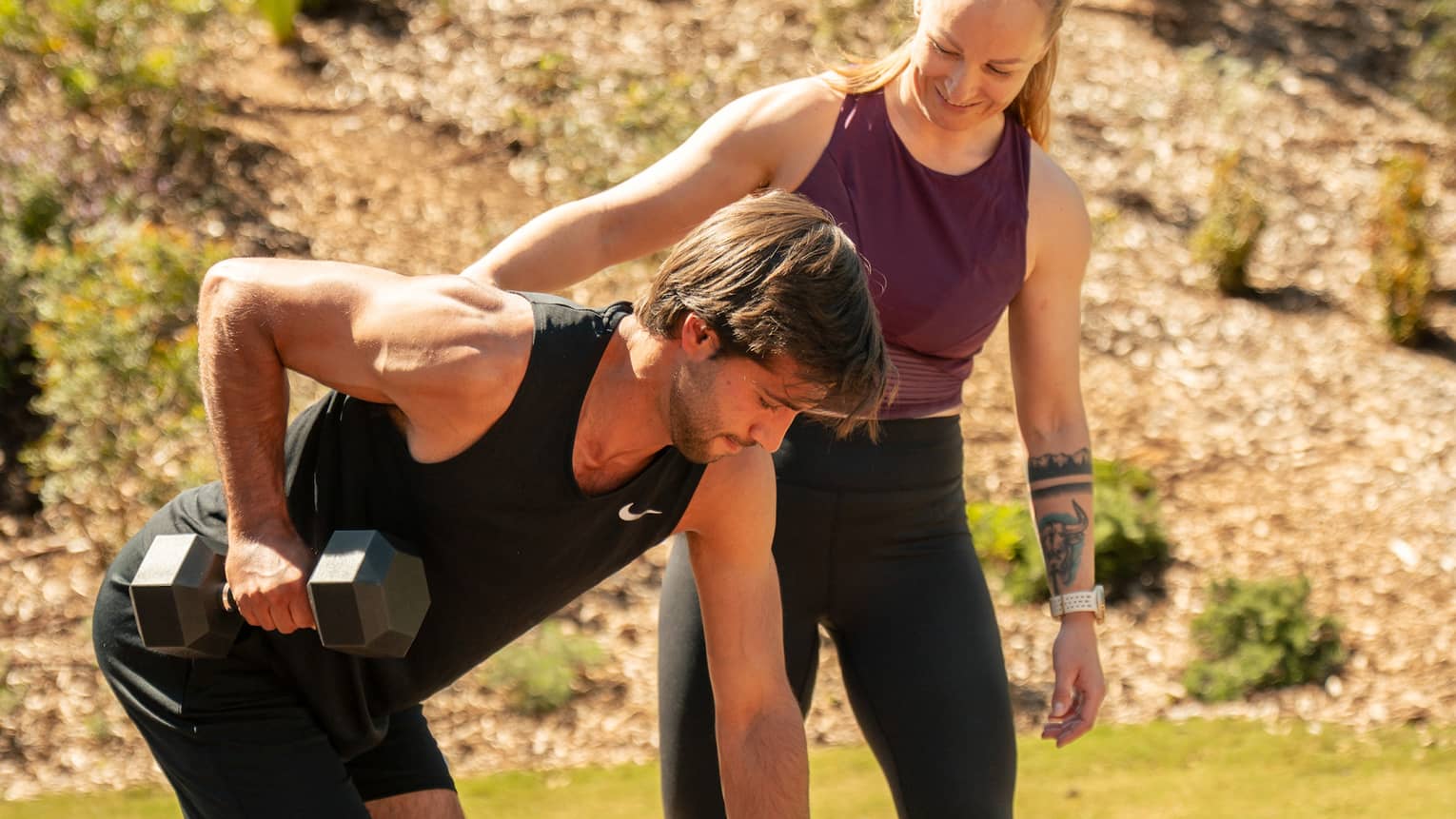 Two people exercise with weights at a workout bench on a wood deck in an outdoor setting.