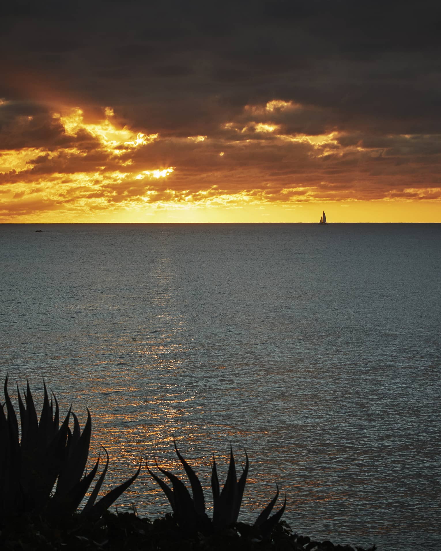 Bright orange sunset over the ocean with dramatic clouds and a sailboat in the distance