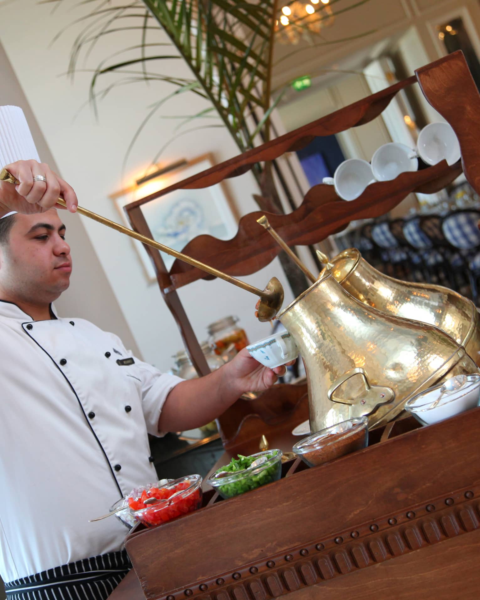Chef in white uniform, tall hat holds long ladle, spoons liquid out of tall brass jug