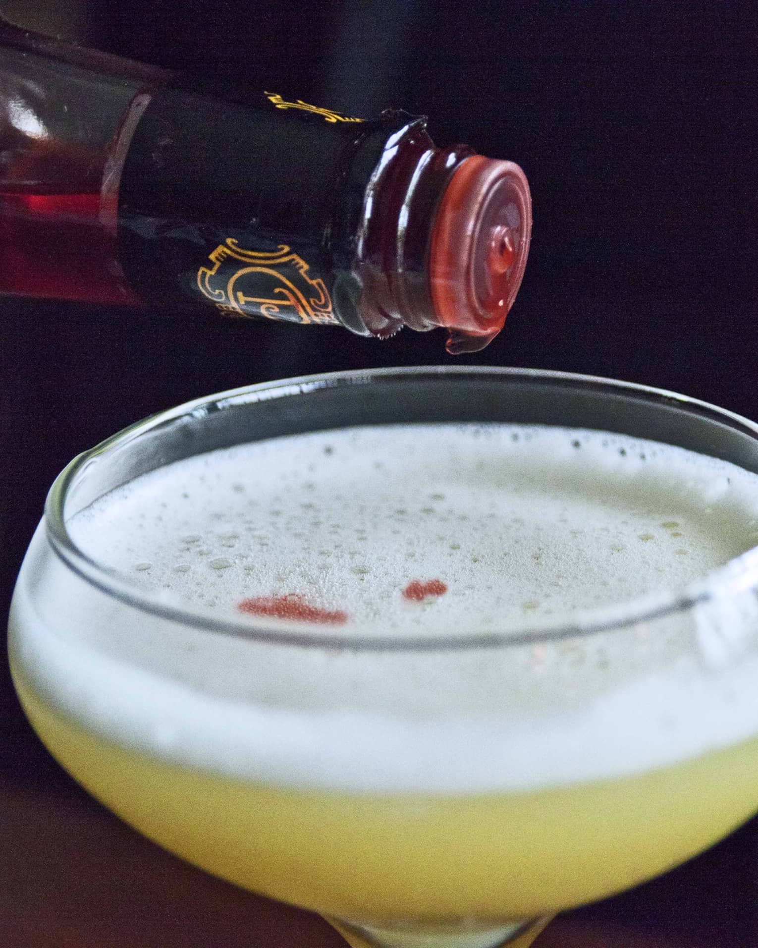 Close-up of martini glass with yellow cocktail, white foam, bottle with red liqueur dripping out