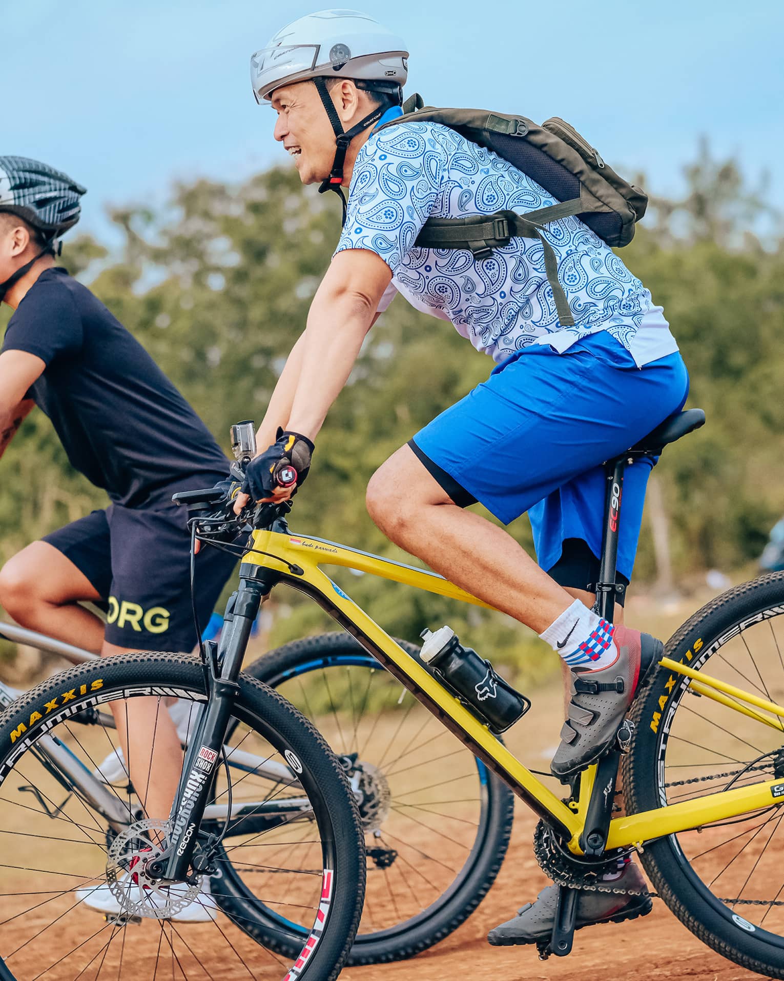 Side view of two smiling mountain bikers in helmets as they coast down a gentle slope on a dirt path alongside trees.