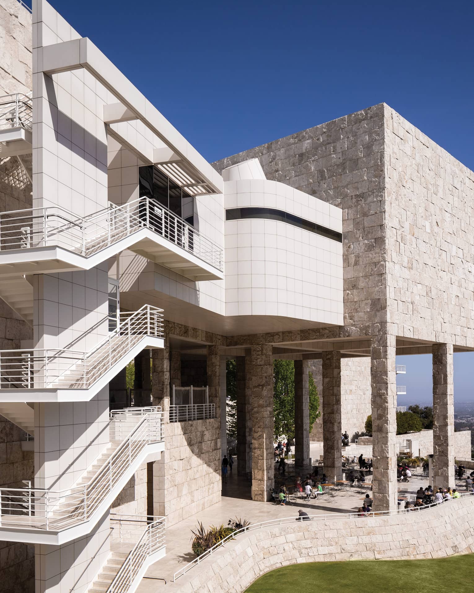 Outdoor stairways line the wall of a tan-coloured stone building with a patio full of people, a city skyline visible beyond.