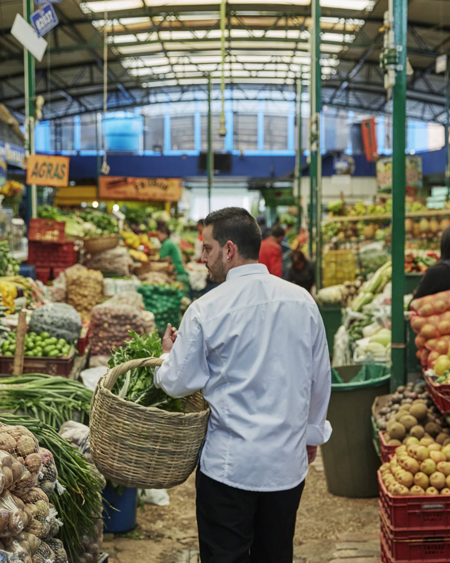 Back of man with beard carrying wicker basket through fruit market aisle, produce stacked on shelves