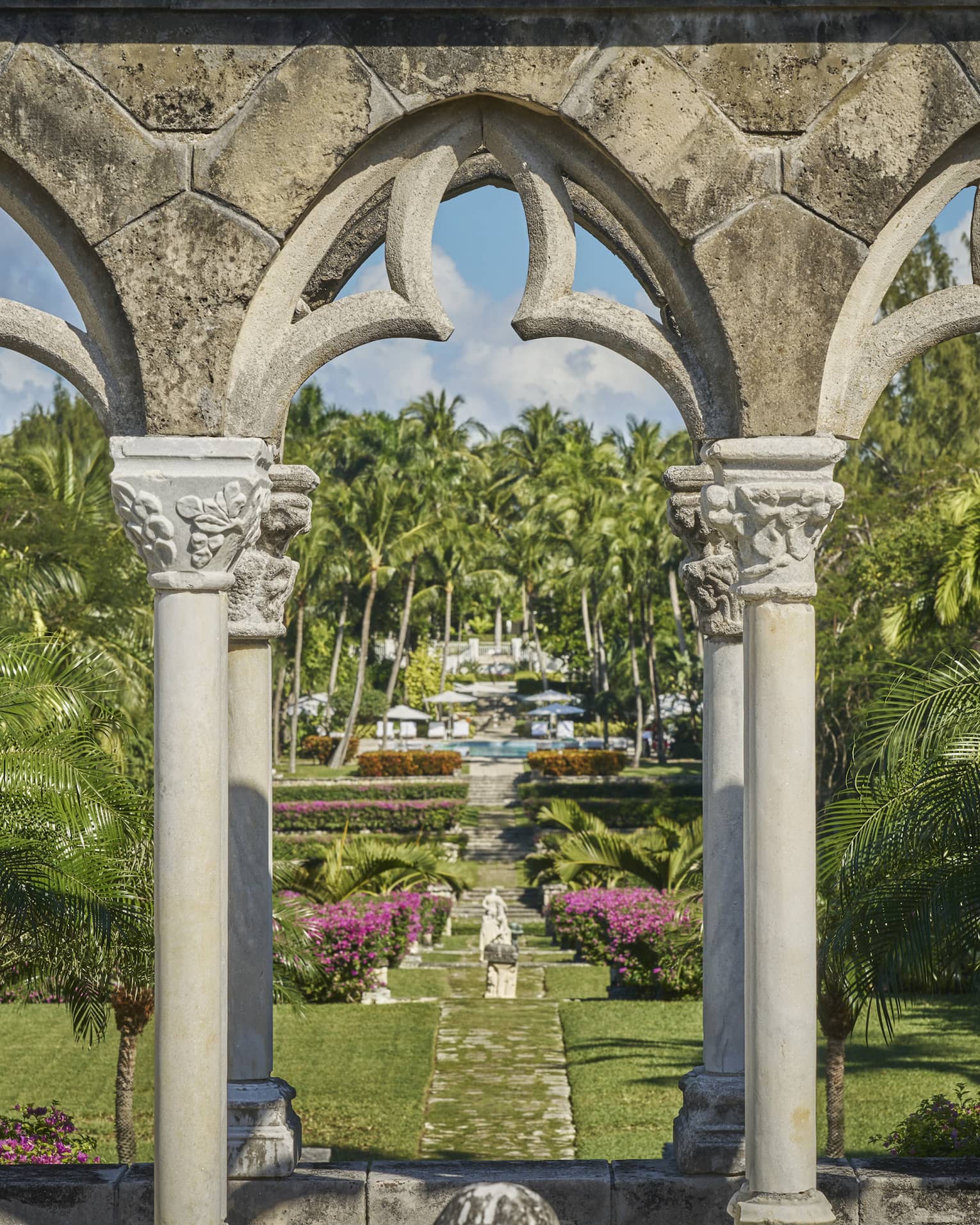 Ornate stone arch leading to a garden pathway with lush greenery, colorful flowers and a statue