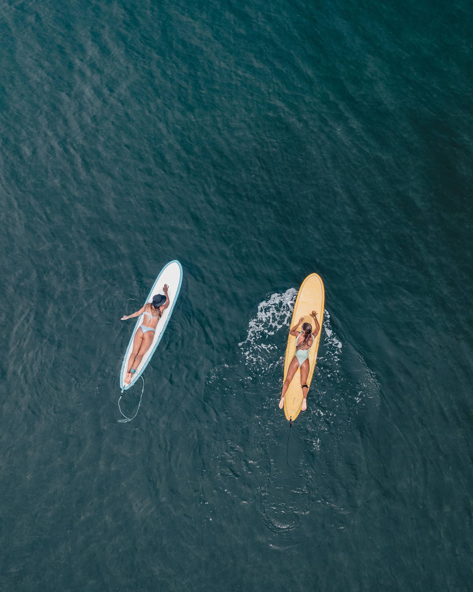 Aerial view of two guests lying on their stomachs on surfboards facing each other on calm water, each wearing a safety leash.