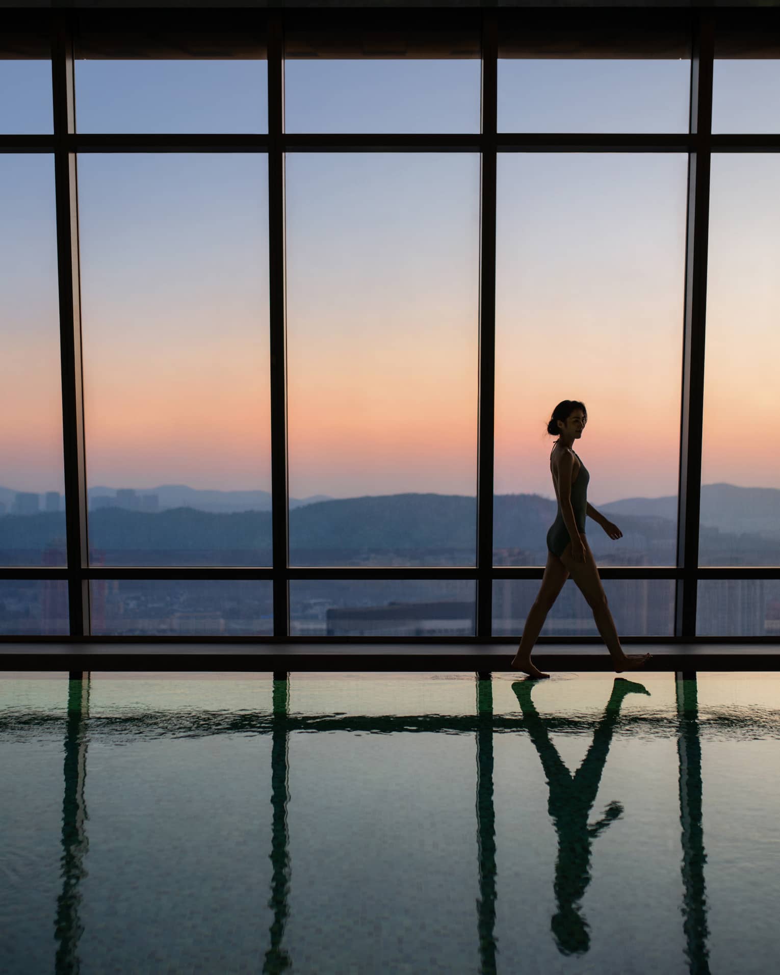 A guest in silhouette walks along the edge of a glassy indoor pool beside a wall of windows with mountain views at sunset.