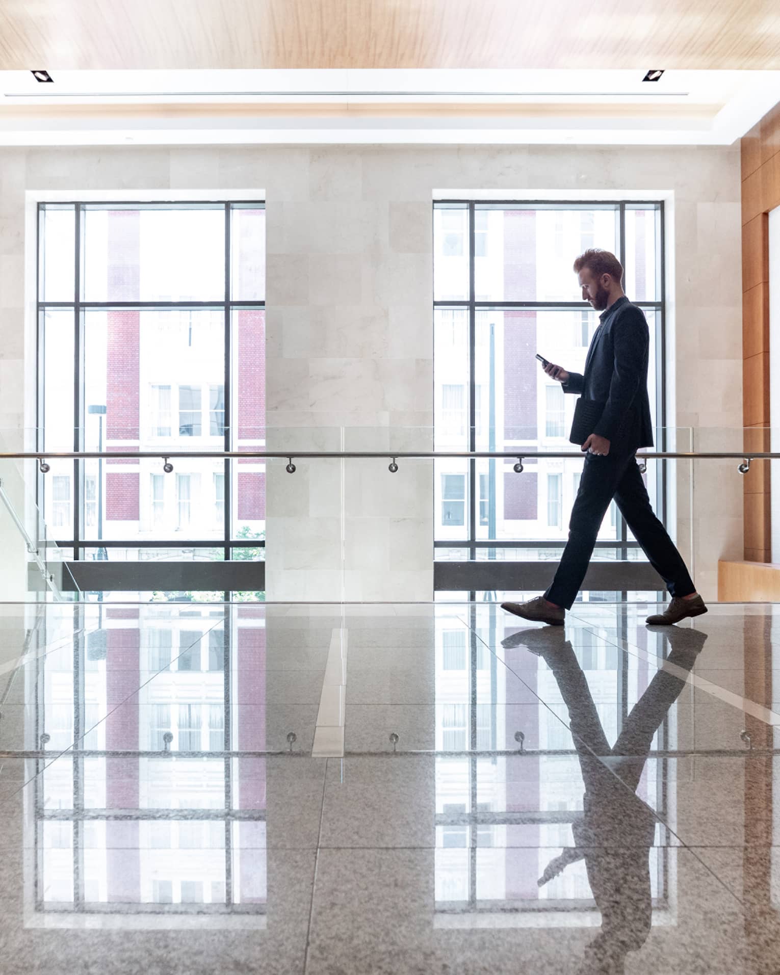 A man looking at his phone while he walks across a lobby area with large windows behind him