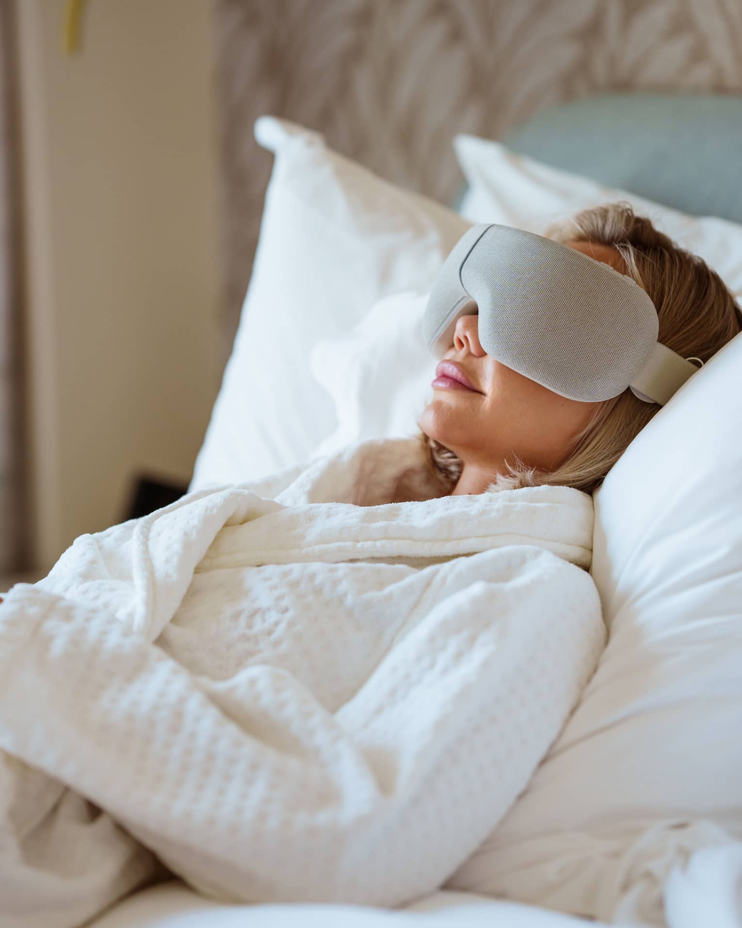 Person wearing white spa robe lays back on a bed with large white pillows wearing a high-tech eye mask