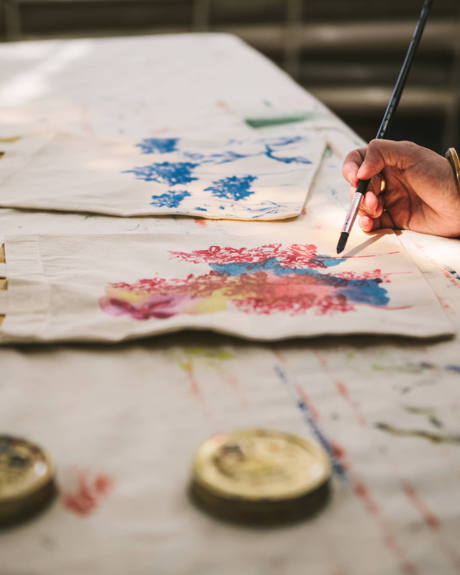 Close-up of a gold-bangled hand wielding a long brush to paint an intricate pattern on a cloth tote bag.