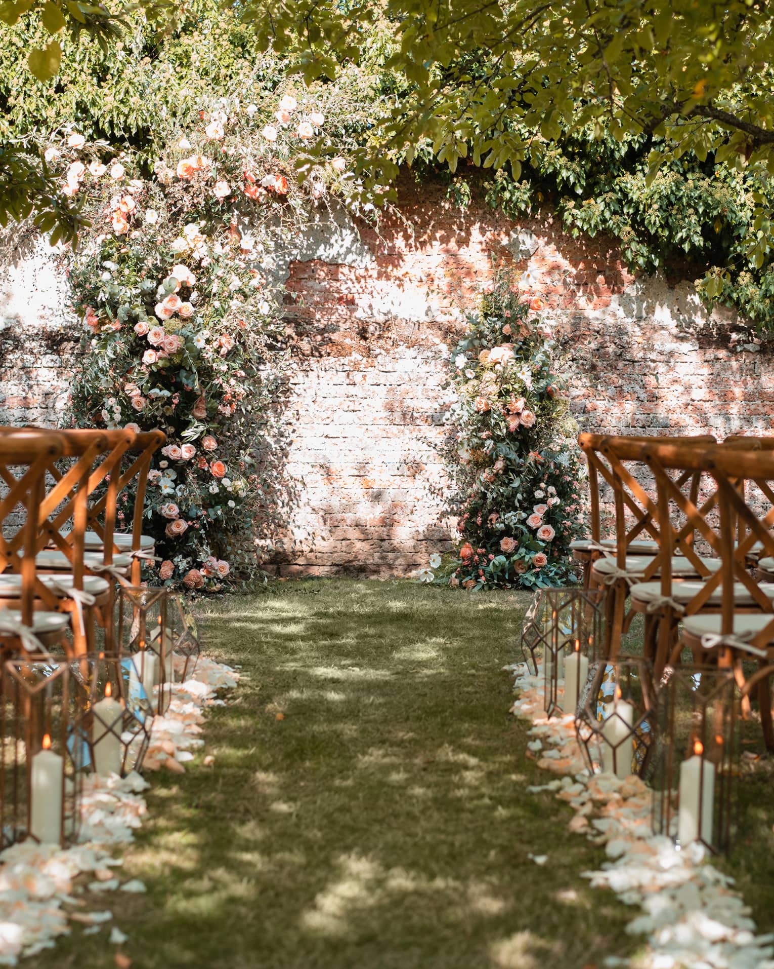 Outdoor wedding aisle lined with flower petals and chairs leading to a brick wall draped with greenery