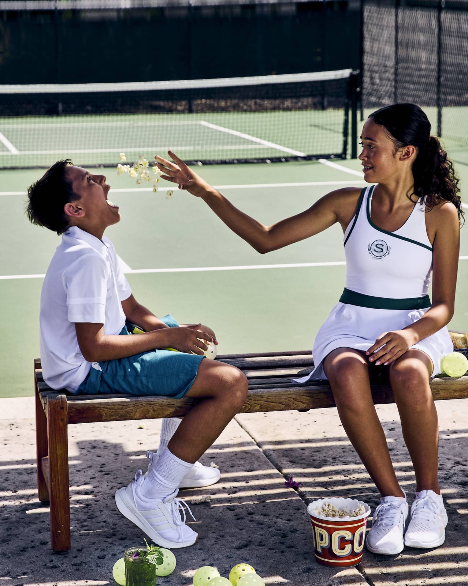 On a shaded wooden bench in front of a pickleball court, two children enjoy some popcorn, pickleballs strewn on the ground.