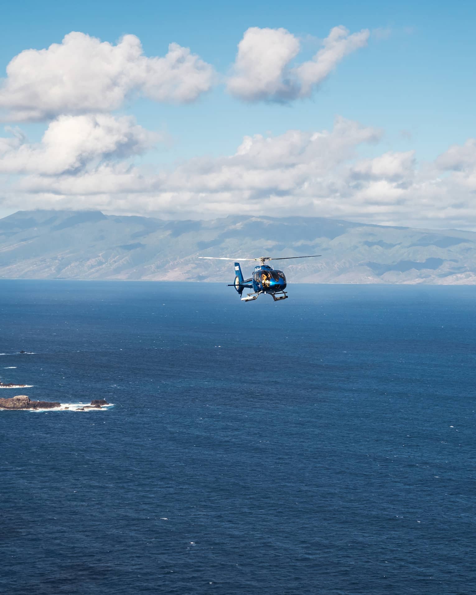 A helicopter flies over open ocean with a mountainous island and blue sky dotted with clouds in the background
