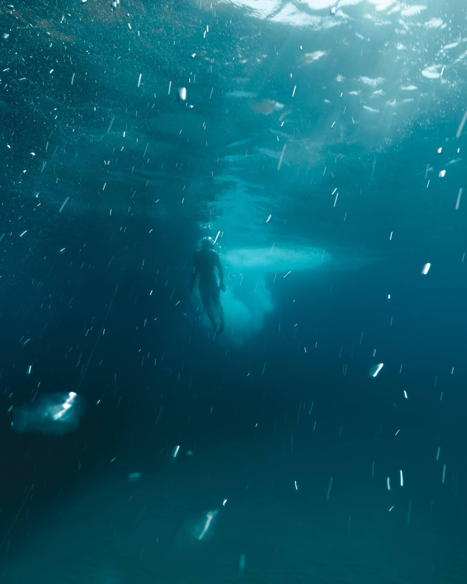 Deep underwater long view of a diver swimming toward the surface as sunlight pierces through the dark blue water.