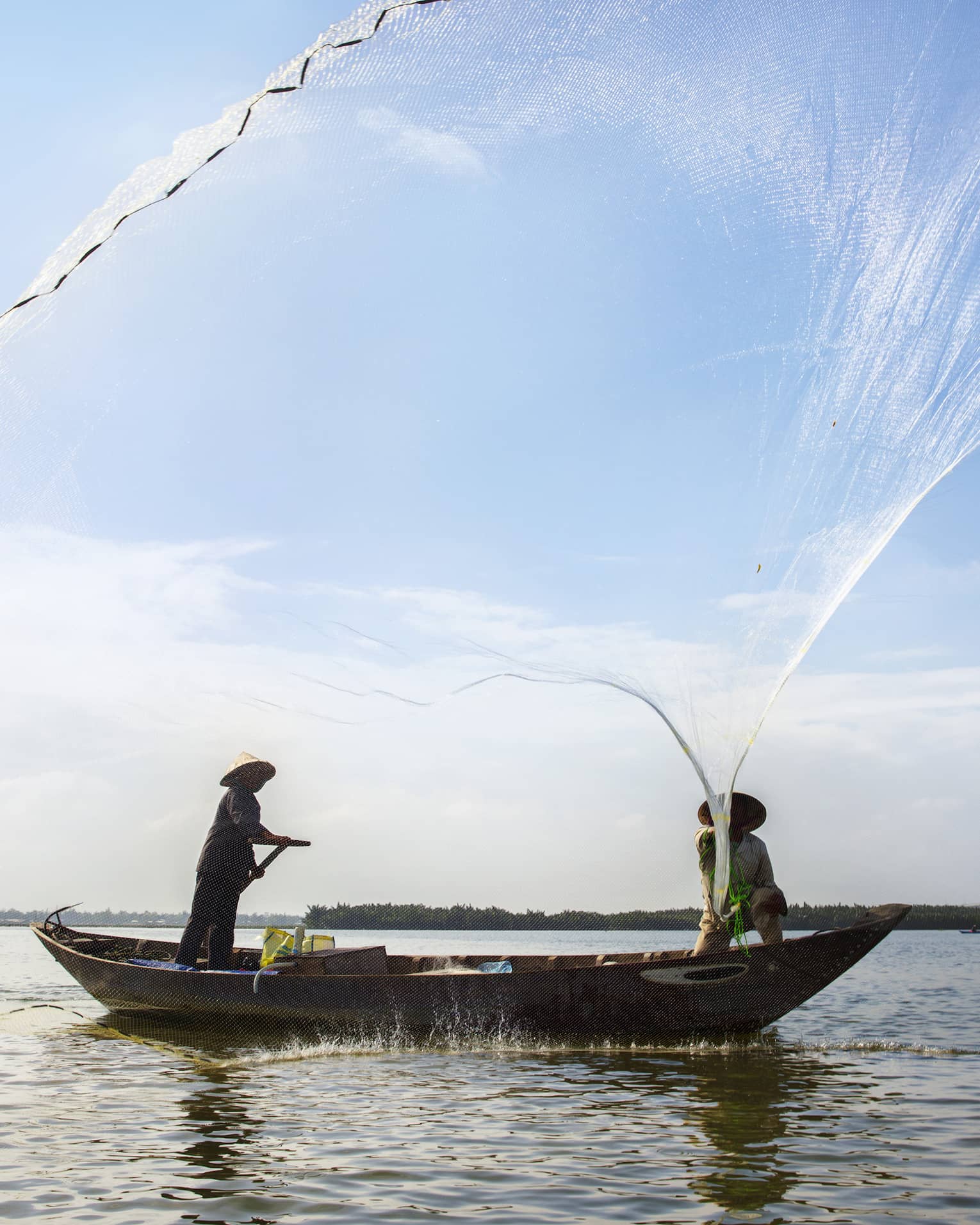 Silhouettes of two people wearing hats in small boat standing, casting large nets 