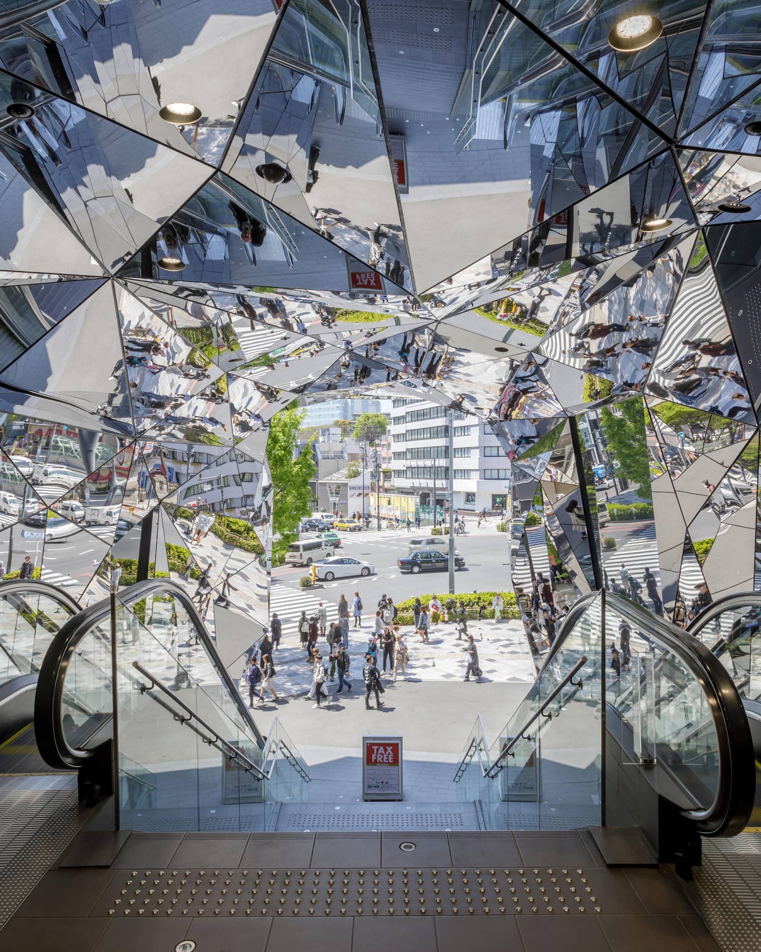 Escalators leading to a mirrored entrance reflecting pedestrians and a busy street scene
