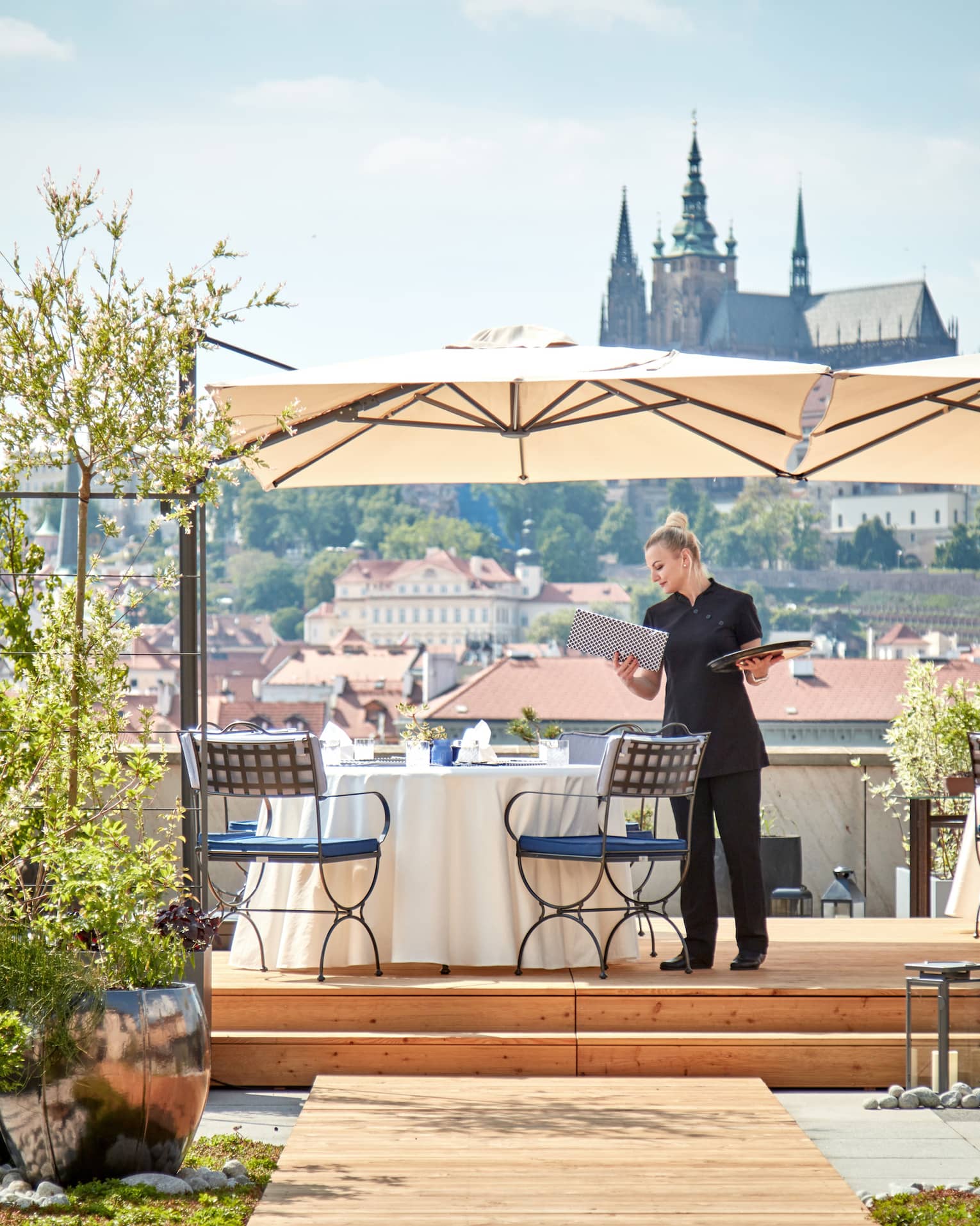 Hotel staff sets dining table under umbrella on rooftop patio overlooking sunny Prague city 