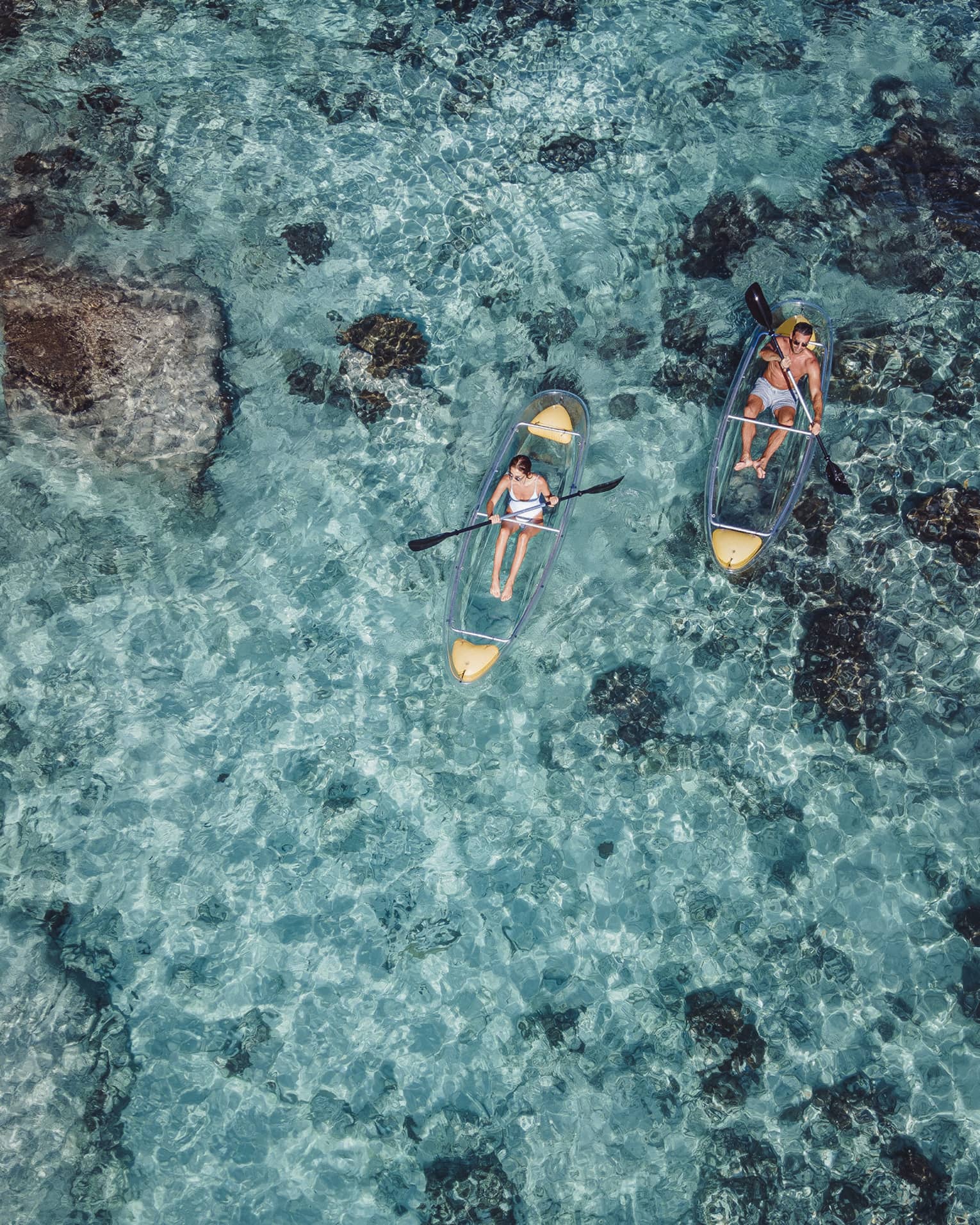 A view from above of kayakers paddling in transparent kayaks near boulders jetting out of crystal-clear open water.