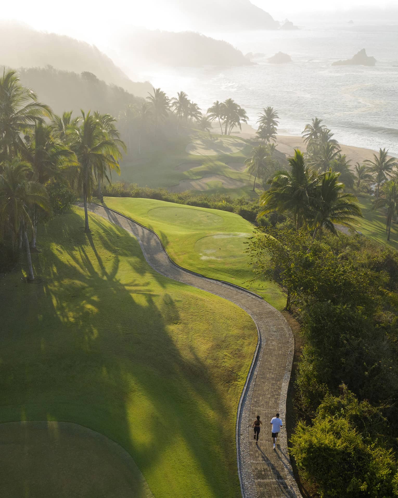 Two people walk along a path among verdant, hilly surroundings, with a beach and shoreline views in the background.