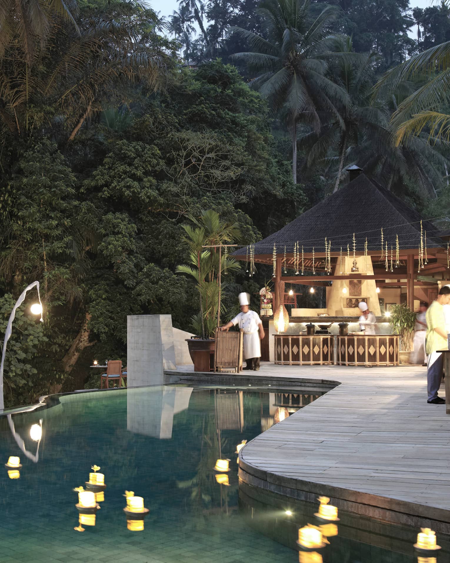 An infinity pool looking out towards the dense jungle; chefs bustle around the outdoor kitchen on the poolside deck.