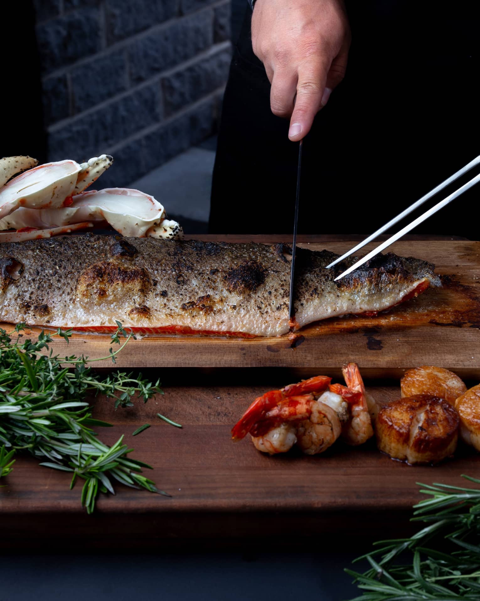 Closeup of chef preparing to carve a side of steelhead trout on a cedar plank, rosemary sprigs and charred lemon garnishes around