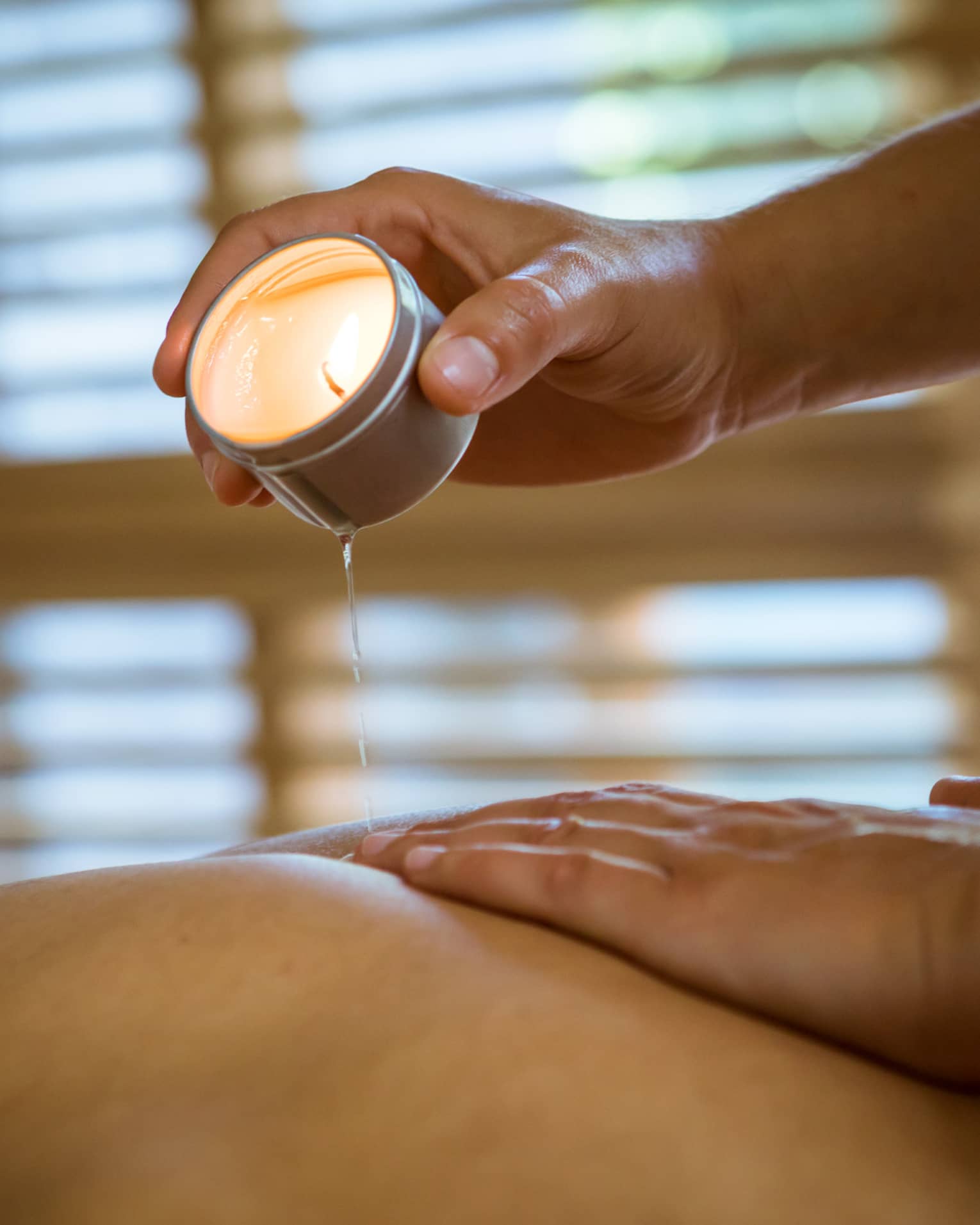 Massage therapist pours hot wax from tealight candle over person's bare back in spa
