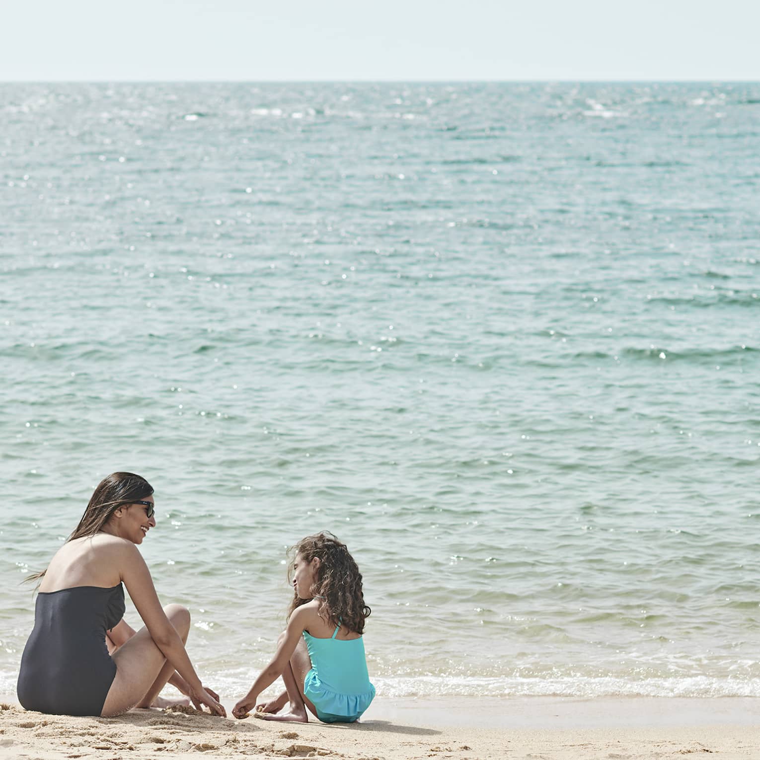 Seated by the water’s edge, a mother and daughter are having fun playing in the sand, while being kissed by the sun’s glow.