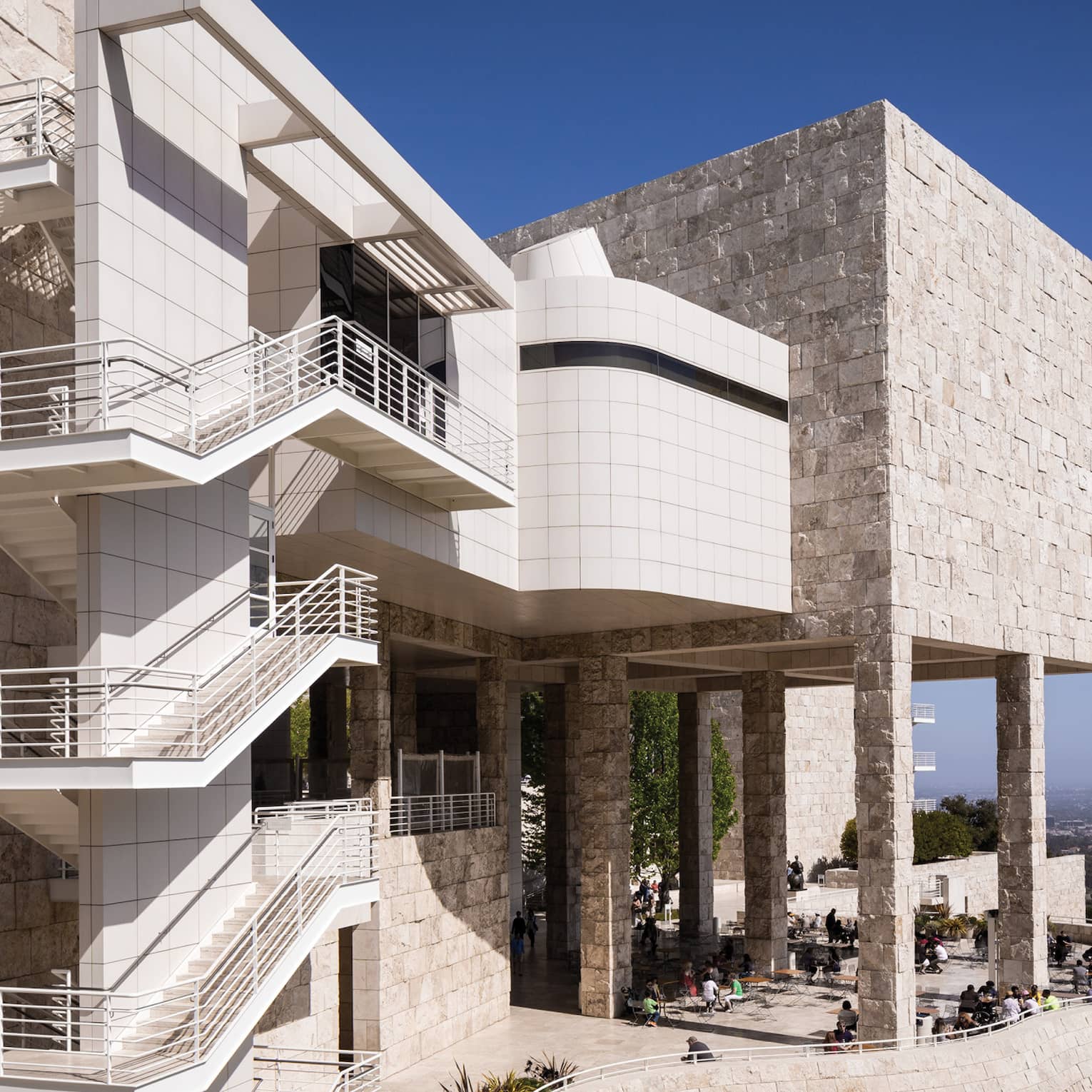 Outdoor stairways line the wall of a tan-coloured stone building with a patio full of people, a city skyline visible beyond.