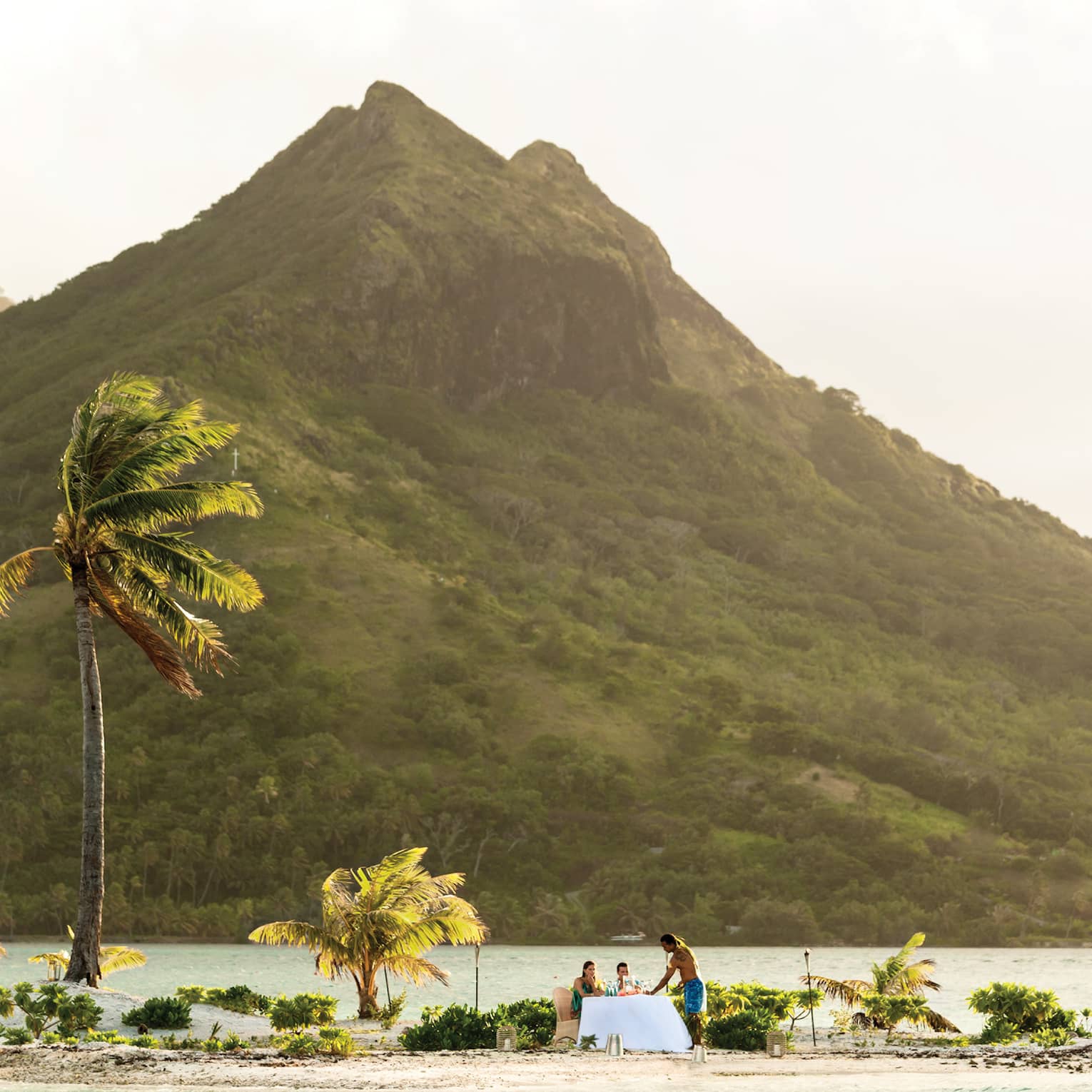 Small tropical island with palm trees, where two people are dining at a table by the water, with a mountain in the background