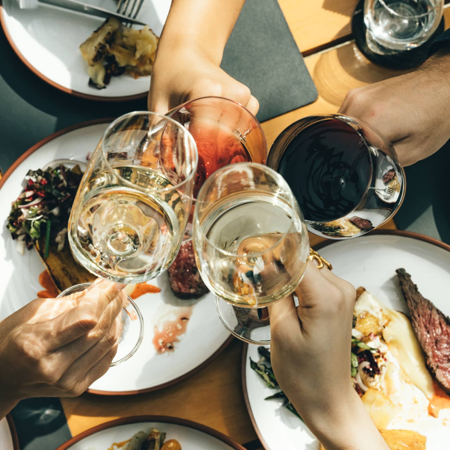 Close-up from above of four hands clinking glasses at the centre of a spread of plates of rare grilled meat and vegetables.