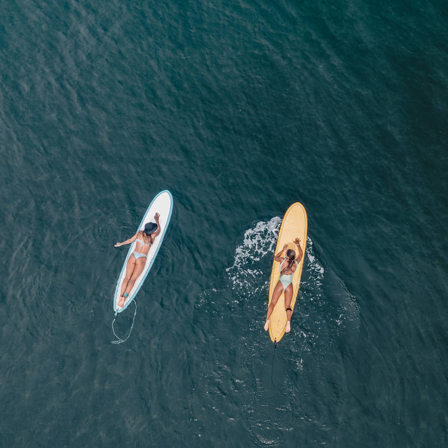 Aerial view of two guests lying on their stomachs on surfboards facing each other on calm water, each wearing a safety leash.