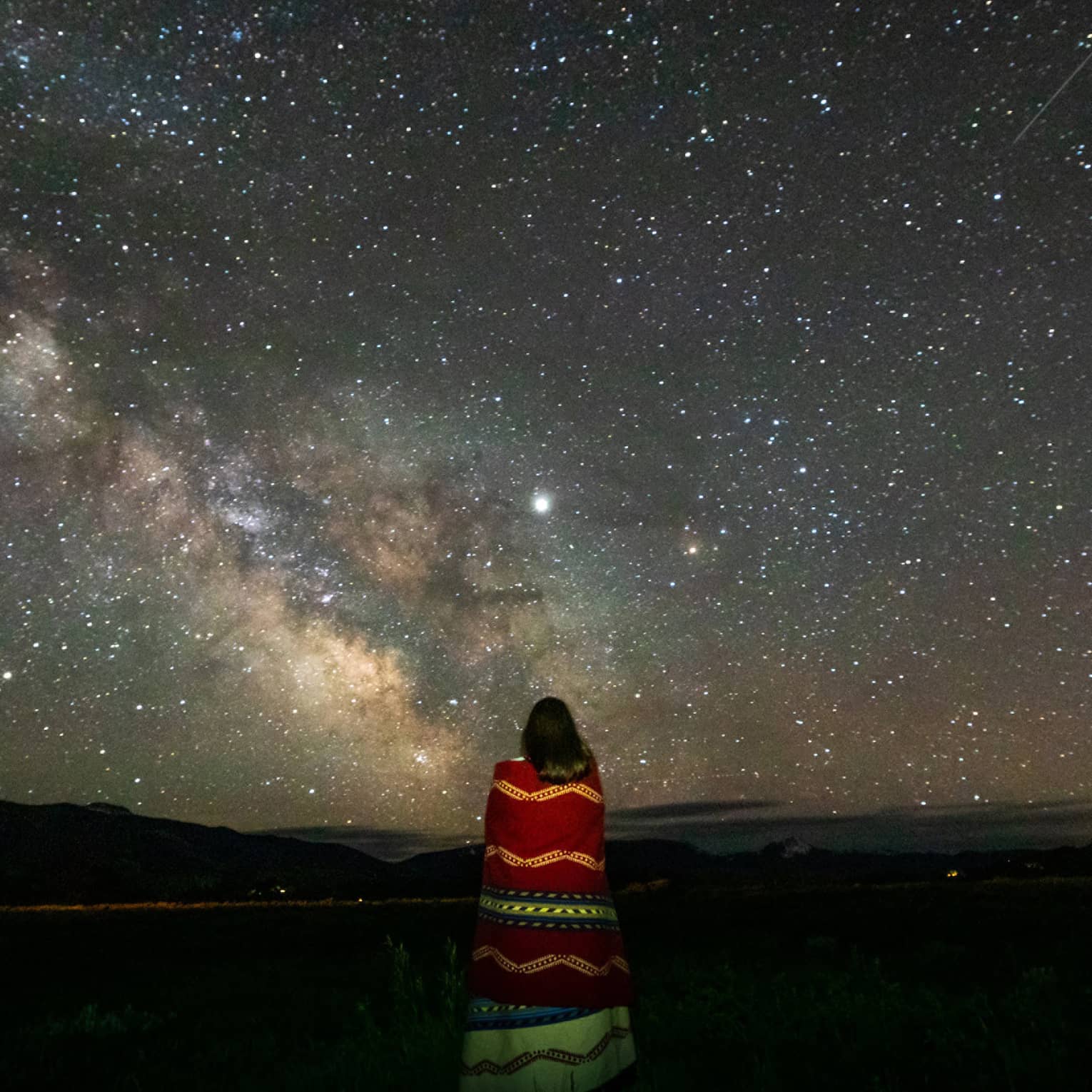 A lone woman stargazes, wrapped in a colourful striped blanket 