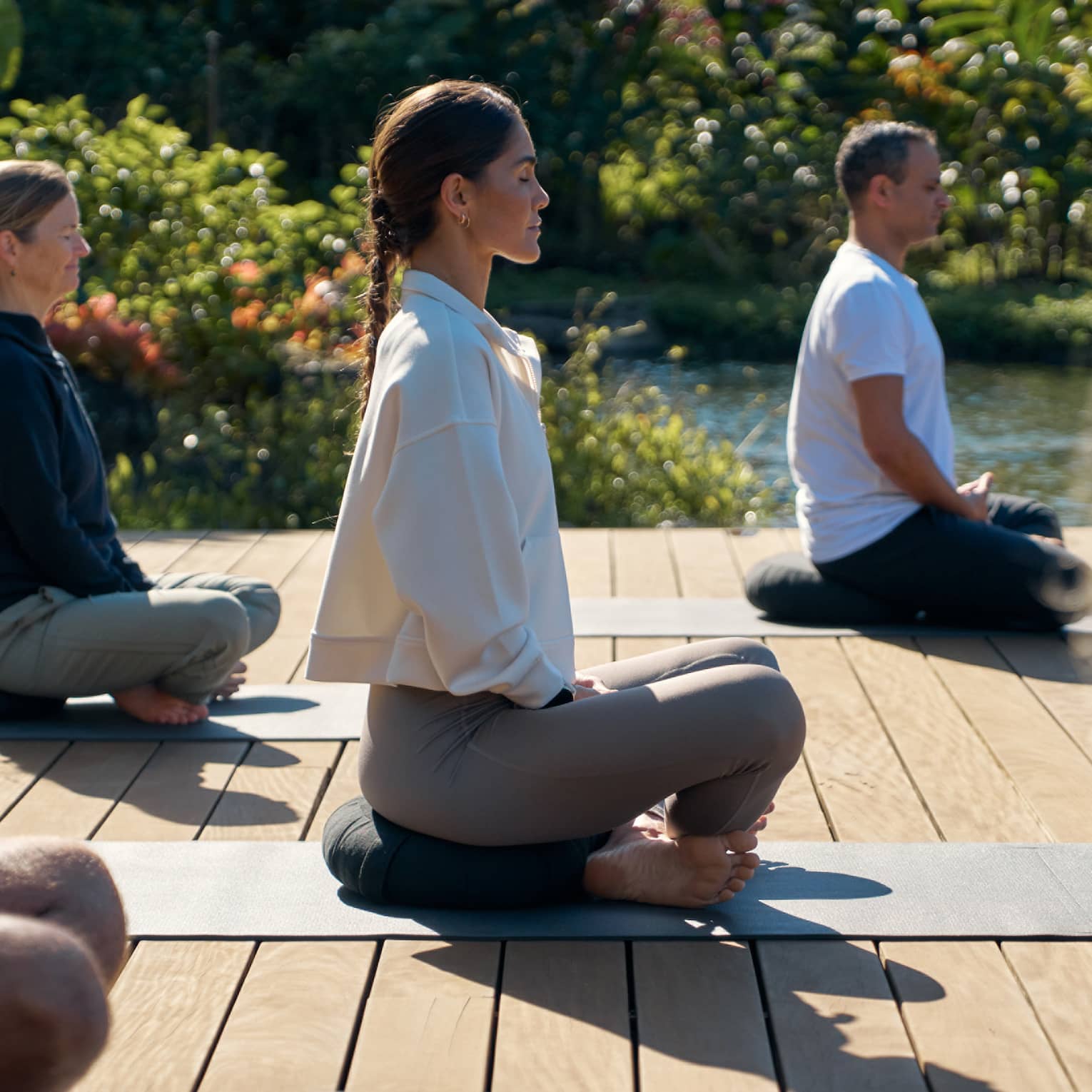A group of people sit cross legged on wooden deck in garden setting, practicing yoga