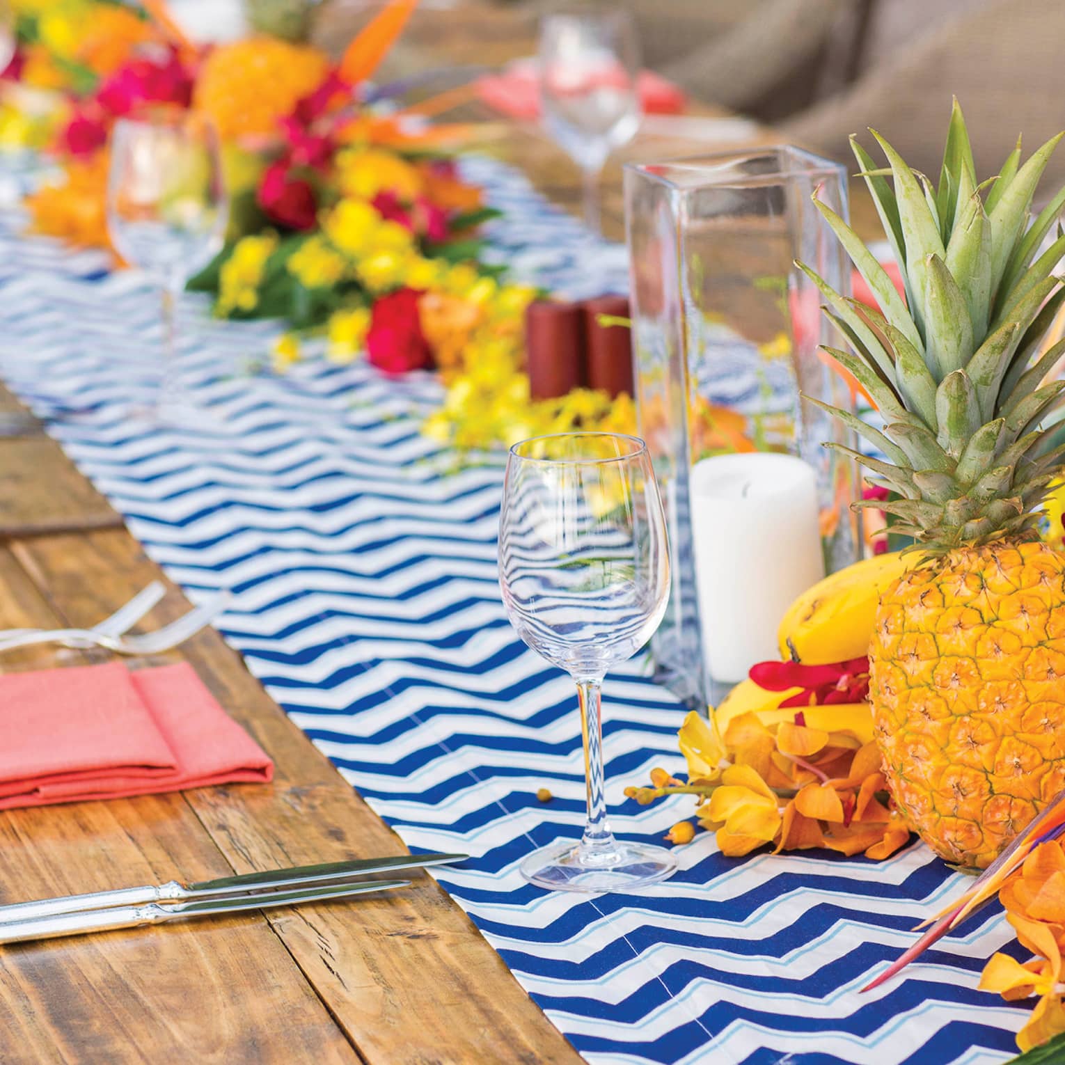 A long orange-hued wooden table set with a blue-and-white zigzag runner, adorned with colourful tropical fruits and flowers.