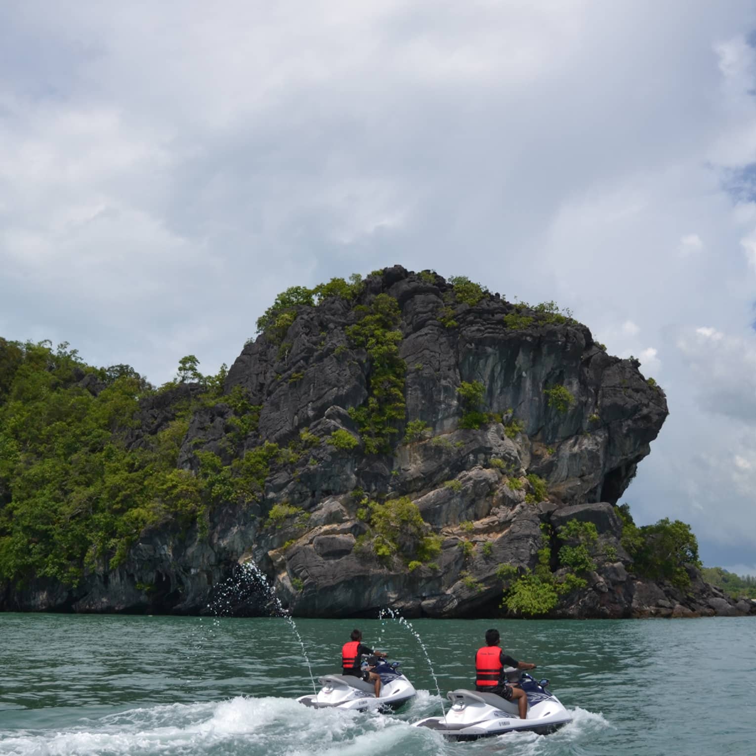 Two people wearing red life vests on jet skis ride on ocean under mountain