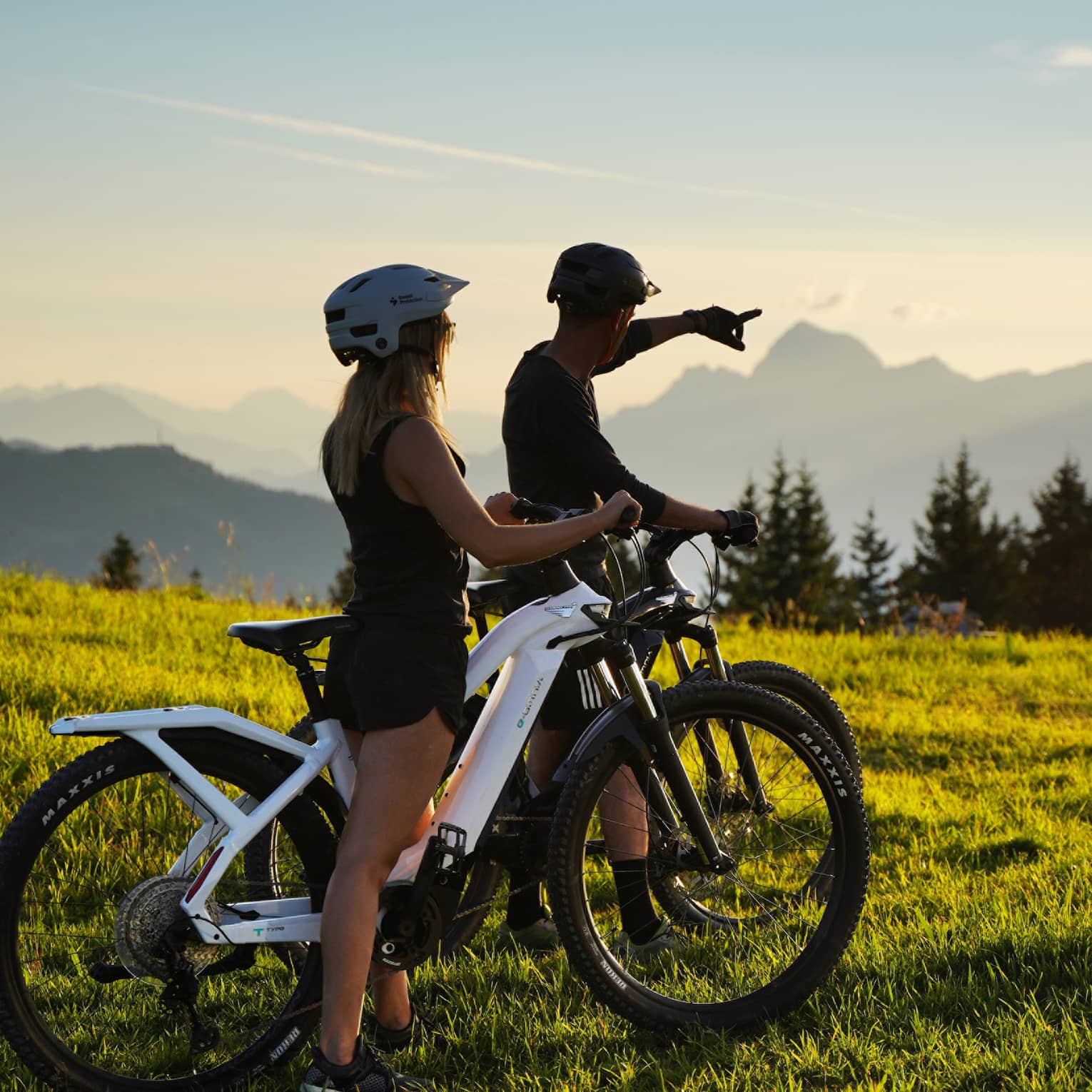 Two people riding fat bikes on meadow, one pointing to distant mountains