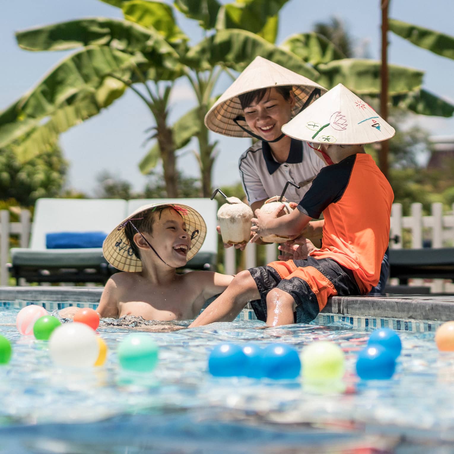 Three laughing children wearing hats, holding coconut drinks play in pool with colourful balls