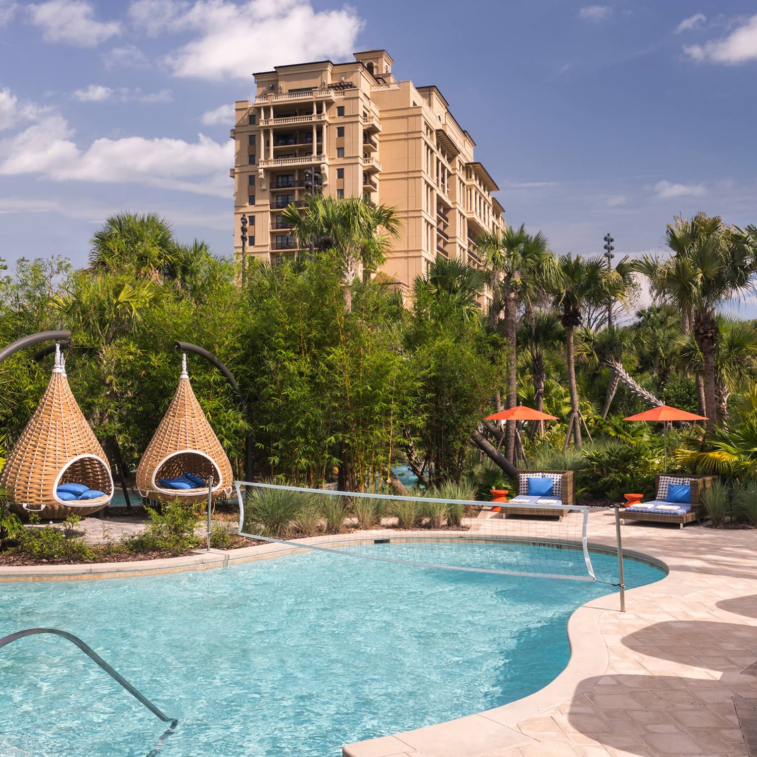 A clear blue pool featuring basketball and volleyball nets is surrounded by palm trees, covered lounges and teardrop daybeds.