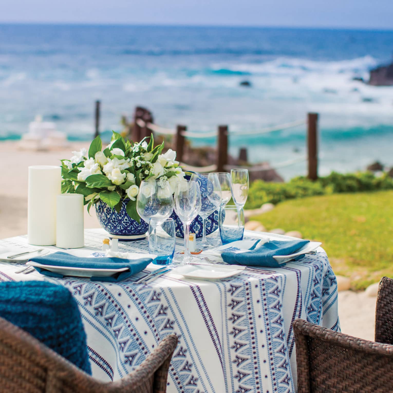 An outdoor table set for two with flowers, blue and white colour scheme, wicker chairs with blue cushions, facing the beach. 