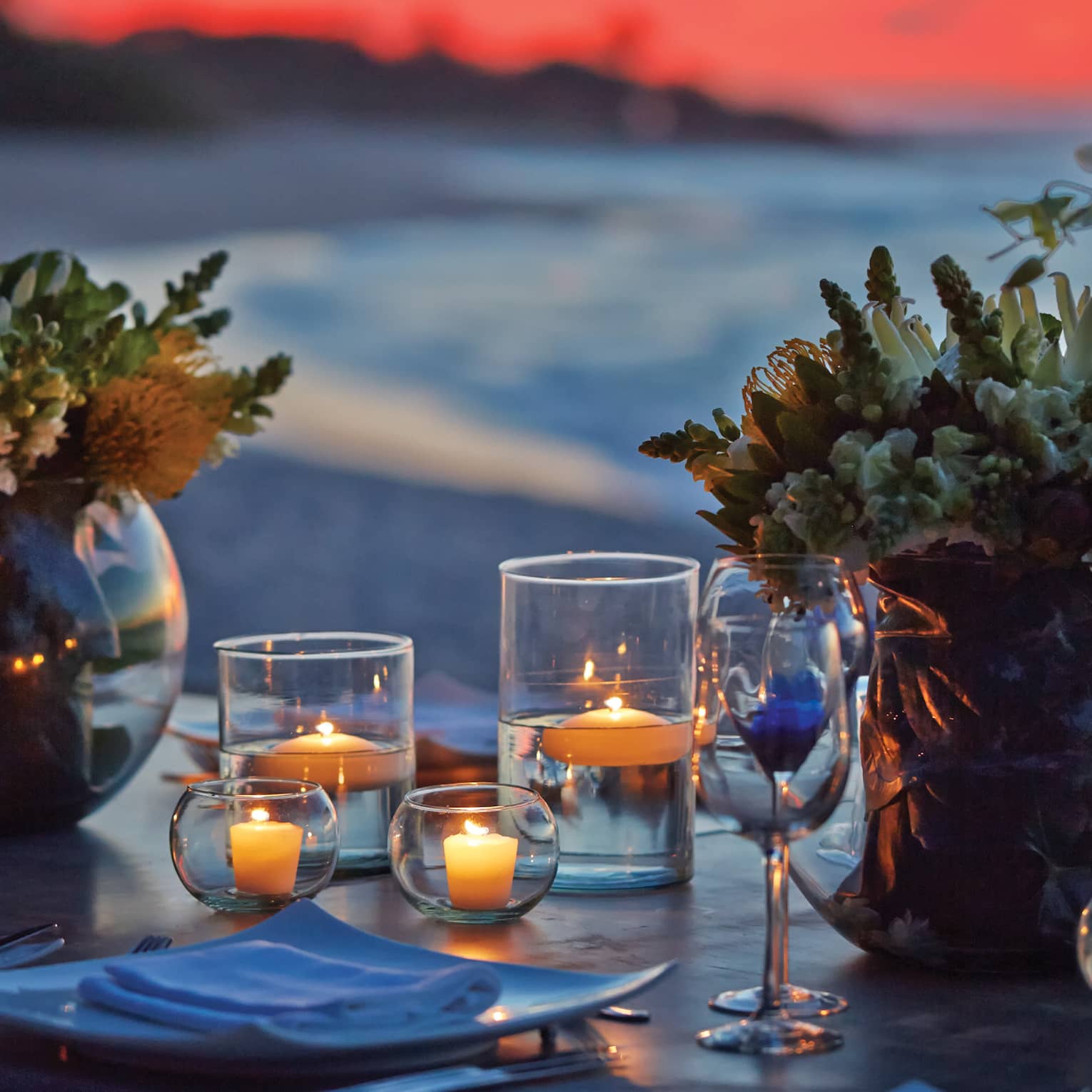 Large round vases of flowers and many small candles in glass holders grace a table facing the beach under a pink-orange sky. 
